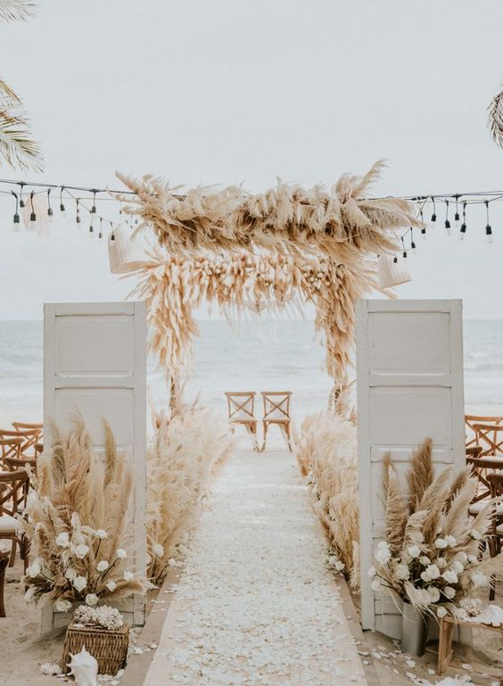 maximalist beach wedding decor with pampas grass, white blooms, simple chairs and white woven lamps