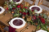 a barn wedding tablescape with an uncovered table, a greenery and burgundy bloom runner, burgundy linens and touches of gold