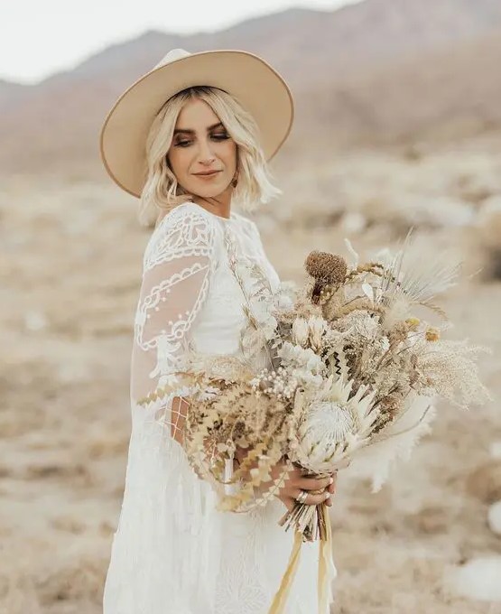 a beautiful boho bridal look with a boho lace wedding dress and a capelet, a neutral hat and a dried flower wedding bouquet for a desert wedding