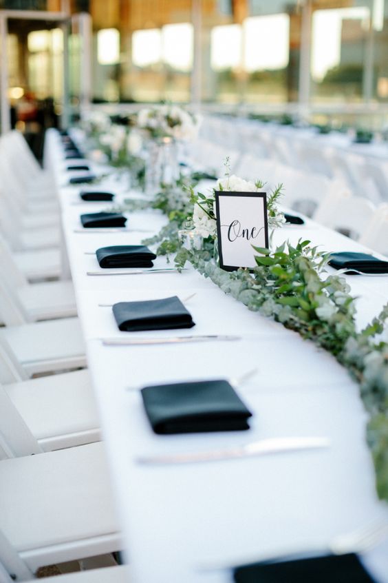 a black and white wedding tablescape with a greenery table runner, black napkins and a black and white table number