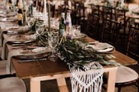 a boho barn wedding tablescape with a macrame and eucalyptus runner, thin and tall candles and hammered metal chargers