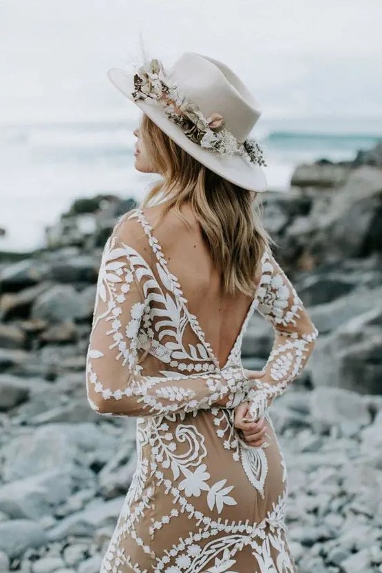 a boho bridal look with a nude and white lace applique wedding dress with long sleeves and a deep cutout back, a white hat with neutral blooms