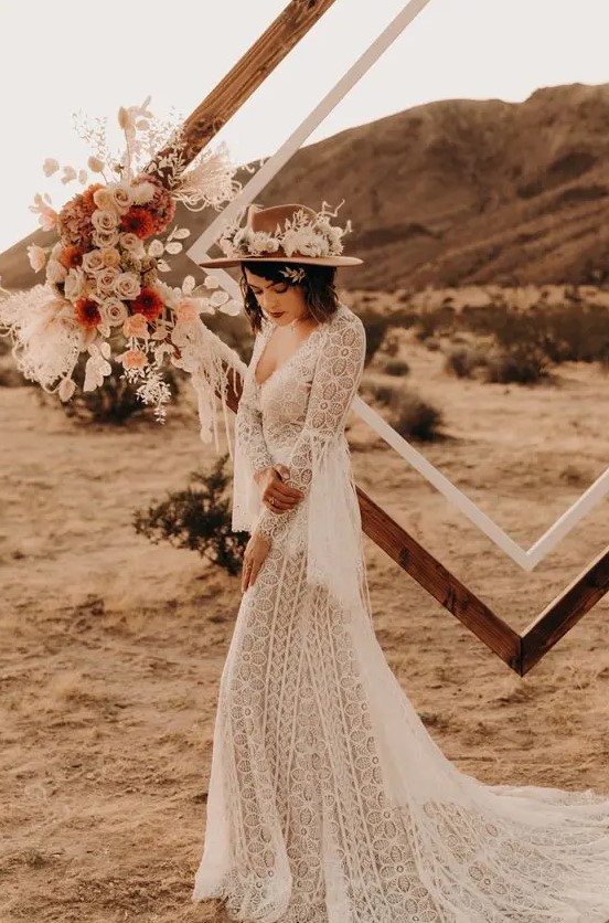 a boho bridal outfit with a boho lace A-line wedding dress with bell sleeves, a depe neckline, a beige hat with white blooms
