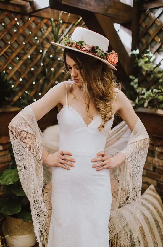 a boho bridal outfit with a lace fitting wedding dress with straps, a boho lace cover up, a white hat with pink blooms and greenery