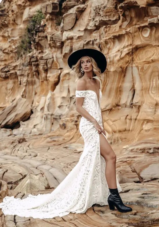 a boho bride wearing a boho lace off the shoulder wedding dress with a plunging neckline and a train, black boots and a black wide brim hat