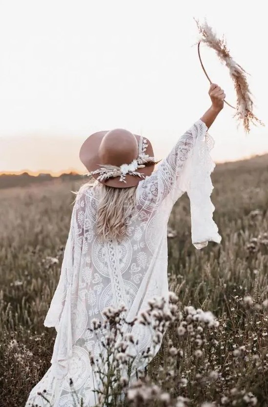 a boho bride wearing a white lace wedding dress with bell sleeves and a brown hat decorated with dried blooms and grass