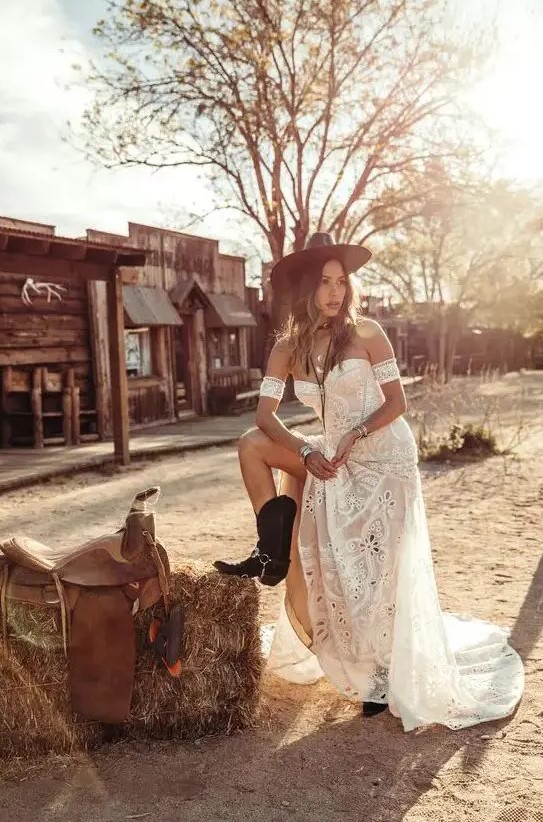 a boho rustic bridal outfit with a strapless boho lace wedding dress, a brown hat and black cowgirl boots, chic boho accessories