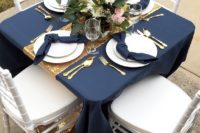 a chic navy and gold table setting with white plates and chargers, gold cutlery and a neutral floral centerpiece