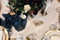 a chic tablescape with much gold, a navy table runner, a moody floral centerpiece and candles