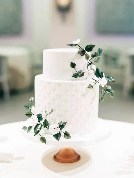 a chic white wedding cake with a plain and geometric tier, with white blooms and green leaves