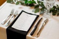 a chic yet simple black and white wedding tablescape with a greenery runner, black napkins and white menus