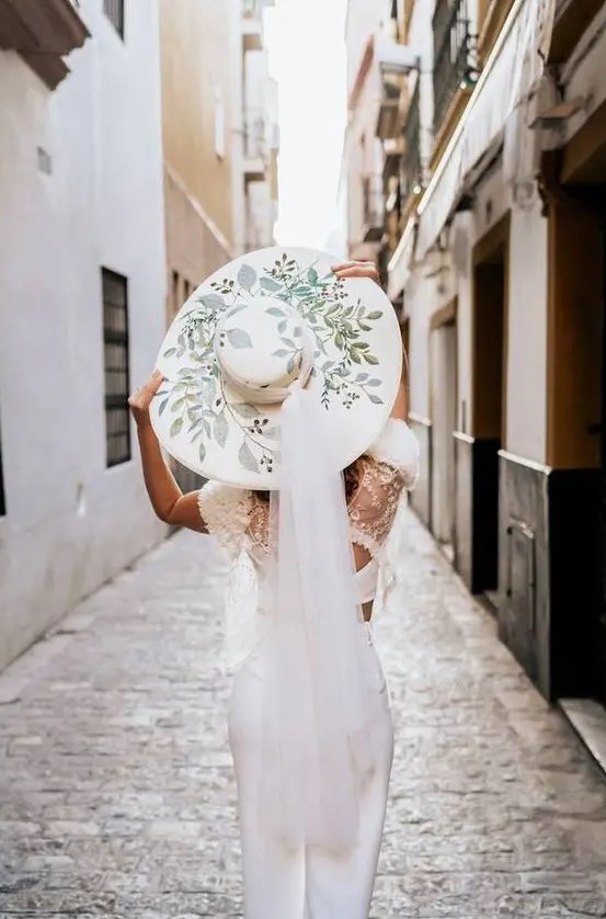 a glam modern bridal look with a white crop top with a lace cover up, white skirt, a wide brim painted hat with a veil