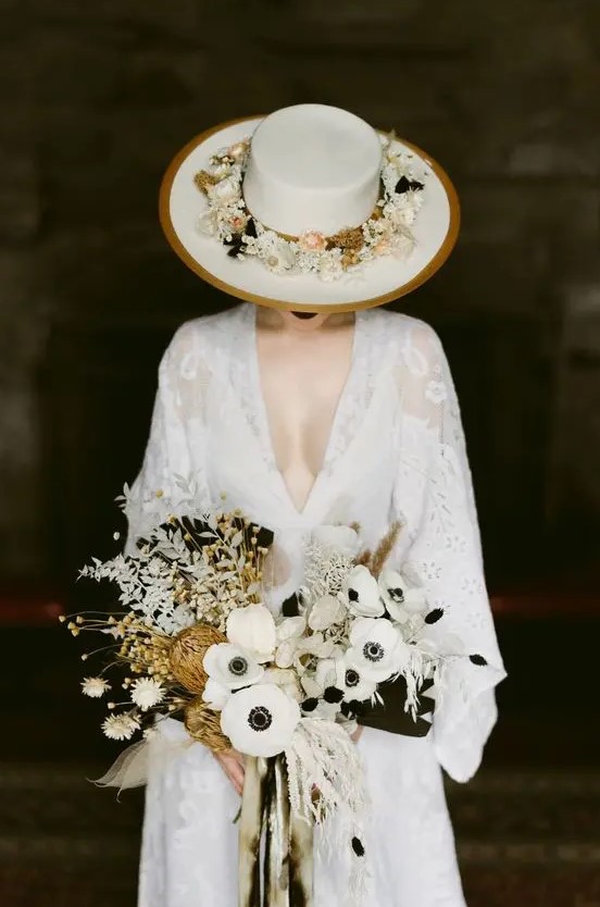a gorgeous boho bridal look with a white lace wedding dress with a plunging neckline, a white hat with gold rims and neutral blooms