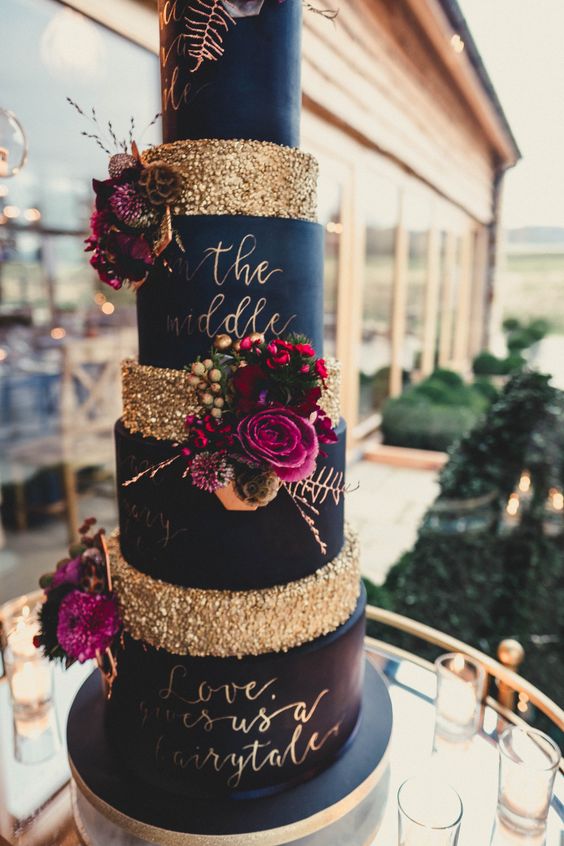 a large and tall navy and gold wedding cake with bright blooms