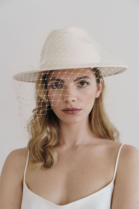 a modern and delicate bridal look with a white hat and a birdcage veil is a chic and lovely idea for a bride