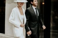a modern bride wearing a white pantsuit with wideleg trousers, a top and an oversized blazer, a white hat with a birdcage veil