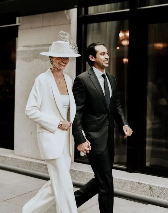 a modern bride wearing a white pantsuit with wideleg trousers, a top and an oversized blazer, a white hat with a birdcage veil