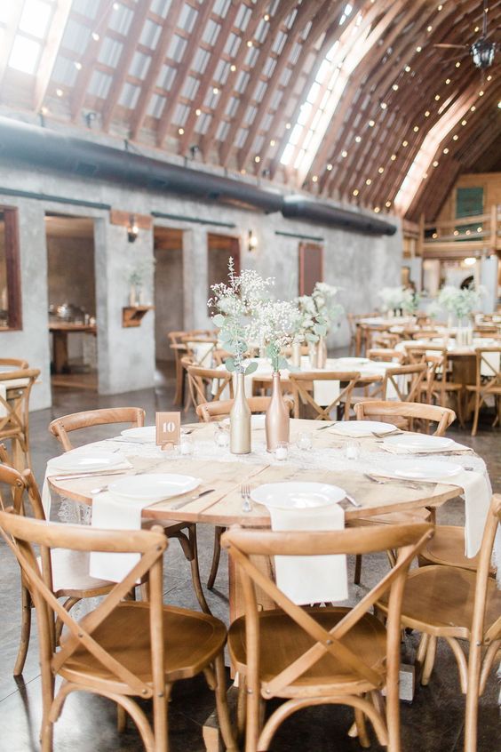 a neutral barn wedding table with neutral linens, metallic bottles with greenery and white blooms and simple cutlery