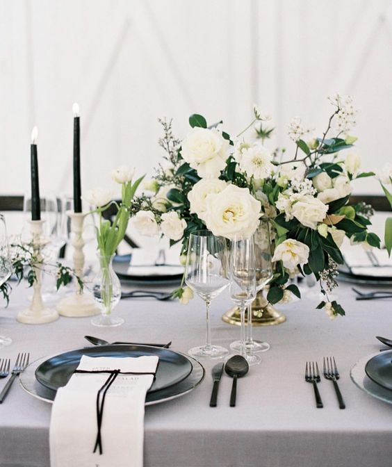 a refined black and white tablescape with a lsuh neutral floral centerpiece, black cutlery, plates and candles plus gold touches