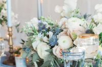 a romantic wedding tablescape with blue candles, glasses and plates plus a neutral floral centerpiece with thistles