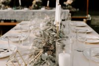 a simple barn-like tablescape with a white tablecloth, a lush eucalyptus runner, candles and terrariums