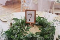 a stylish barn wedding table with white linens, a lush greenery centerpiece and a table number
