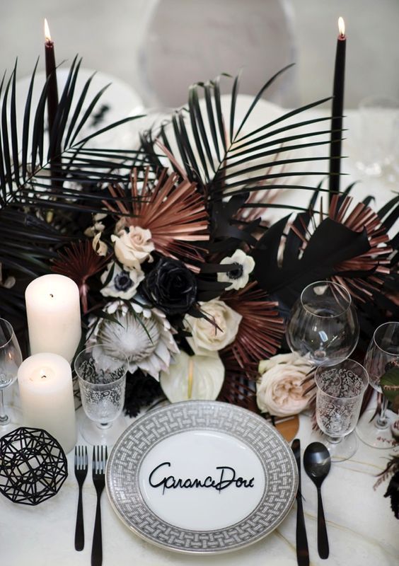 a stylish modern tropical tablescape with black and pink fronds, black candles and cutlery and white pillar candles