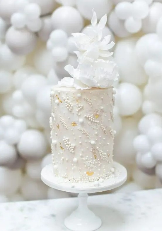 a tall neutral wedding cake decorated with edible pearls and beads and with white sugar blooms on top