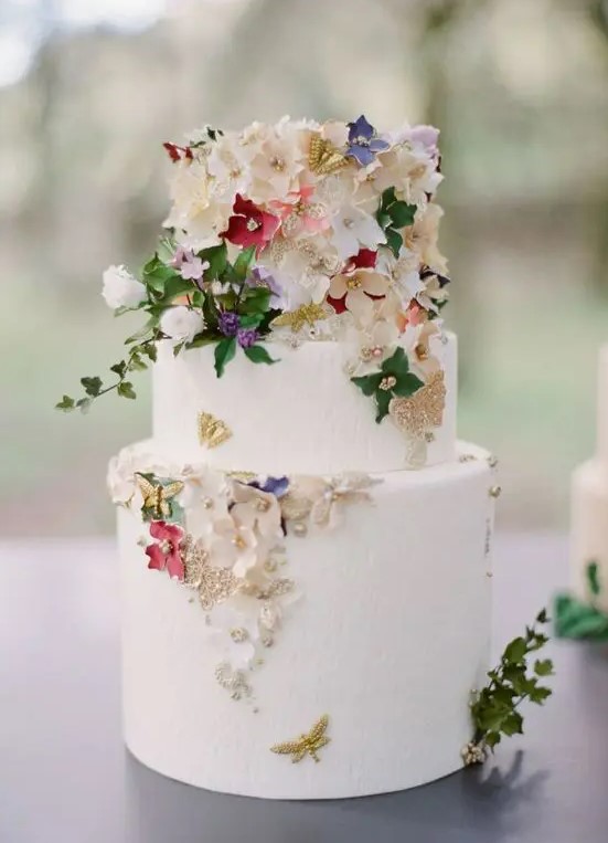 a white buttercream wedding cake decorated with gold foil, sugar flowers and real leaves, beads and butterflies is amazing