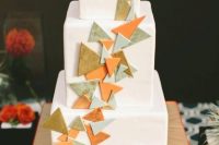 a white square wedding cake with colorful 3D triangles decorating it and an orange topper with a heart is cool and fun