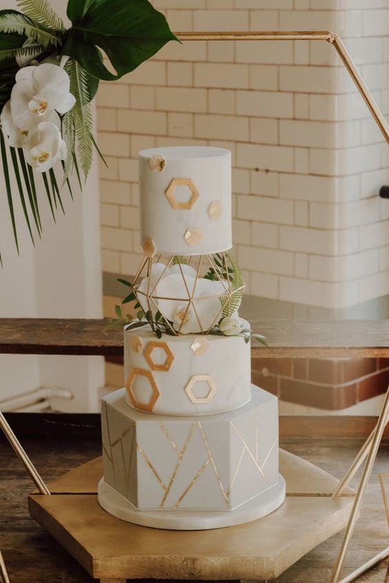 a white wedding cake with gold hexagons and geometric patterns, white orchids and greenery is a chic and refined idea