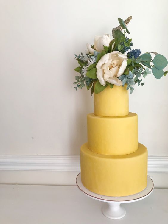 a yellow wedding cake with greenery and neutral blooms on top is a chic and bold idea for a spring or summer wedding