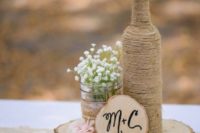 a barn wedding centerpiece of a wood slice, a bottle wrapped with twine, a jar with baby’s breath, a pink rose and a monogram on a wood slice
