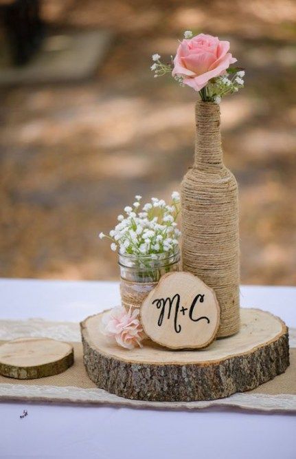 a barn wedding centerpiece of a wood slice, a bottle wrapped with twine, a jar with baby's breath, a pink rose and a monogram on a wood slice