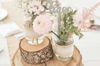 a barn wedding centerpiece of wood slices, jars wrapped with burlap and lace, neutral blooms and greenery and a calligraphy table number