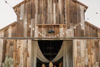 a barn wedding ceremony space with white florals and greenery arrangements, candle lanterns and potted trees plus a fabric decoration