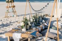 a beach wedding picnic with lights, a low table, lots of pillows, greenery and white blooms, neutral candles