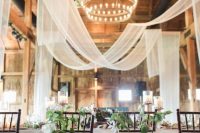 a beautiful barn wedding venue with neutral fabric, blooming branches and a tiered chandelier in the center of the venue