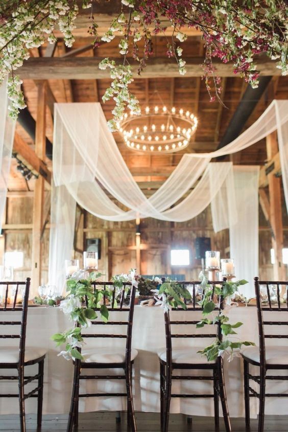 a beautiful barn wedding venue with neutral fabric, blooming branches and a tiered chandelier in the center of the venue