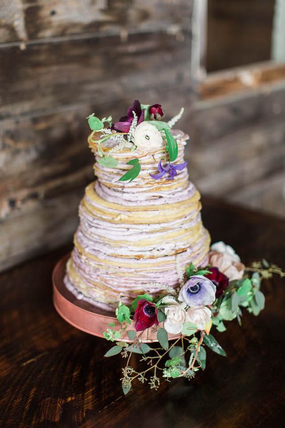 a beautiful crepe wedding cake with lavender cream, white, lilac, burgundy blooms and greenery and fruit slices is chic