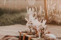 a boho wedding picnic setting with a large chandelier, pampas grass, candles, pillows and a white leather ottoman