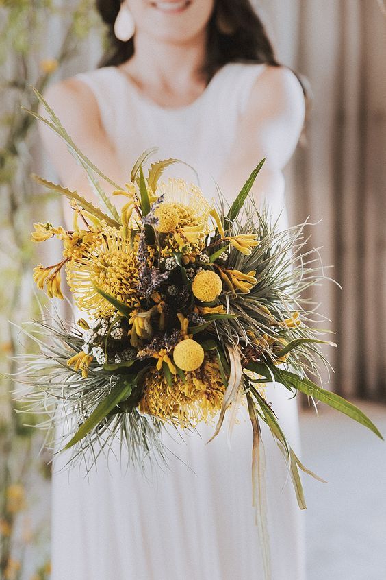 a bold textural wedding bouquet of greenery, pincushion proteas, billy balls and other yellow blooms for a spring or summer bride