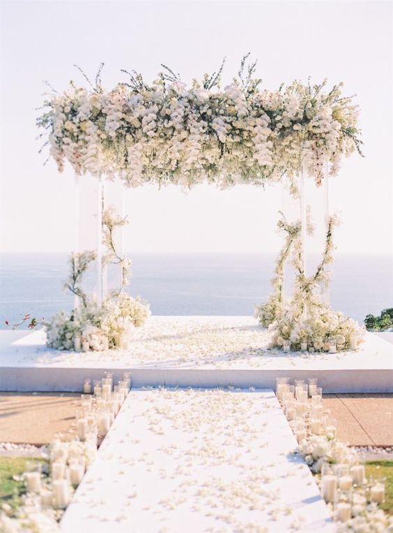 a breathtaking wedding ceremony space with a sea view, an acrylic arch with lush white florals, pillar candles lining up the aisle