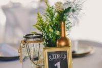 a chic and simpel barn wedding centerpiece of a wood slice, a table number, greenery and neutral blooms, a gilded bottles and a candle lantern