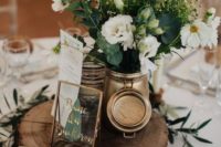 a chic barn wedding centerpiece of a wood slice, greenery, a gilded jar with neutral blooms and greenery and a metallic table name