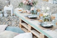 a chic coastal bridal shower picnic with a pallet table, lots of pillows, pastel blooms, gold glasses and blue plates plus greenery