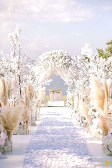 a chic wedding ceremony space with pampas grass and white blooming branches lining up the aisle and covering the arch, with petals on the floor