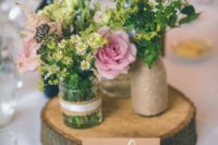 a cozy barn wedding centerpiece of a wood slice, a vase wrapped with twine, jars with lace and wildflowers