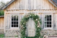 a cozy outdoor barn wedding ceremony space with a greenery and floral arch, some benches with florals and fabric covers
