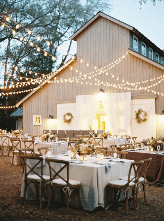 a cozy outdoor barn wedding reception space with elegant blue linens, stained chairs, white florals and greenery and string lights